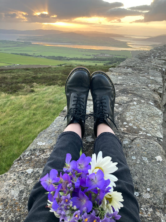 A sunset view from Grianan of Aileach hill fort in Donegal, Ireland. In the foreground flowers are sitting on someone's legs as they sit on the wall of the fort.